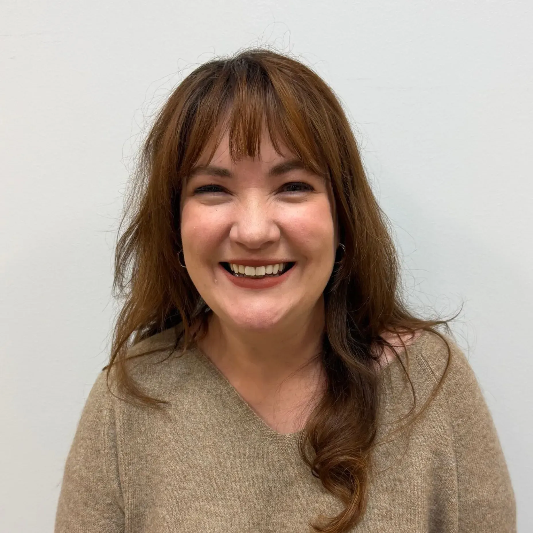 Woman with brown hair and bangs smiles, wearing a tan sweater.