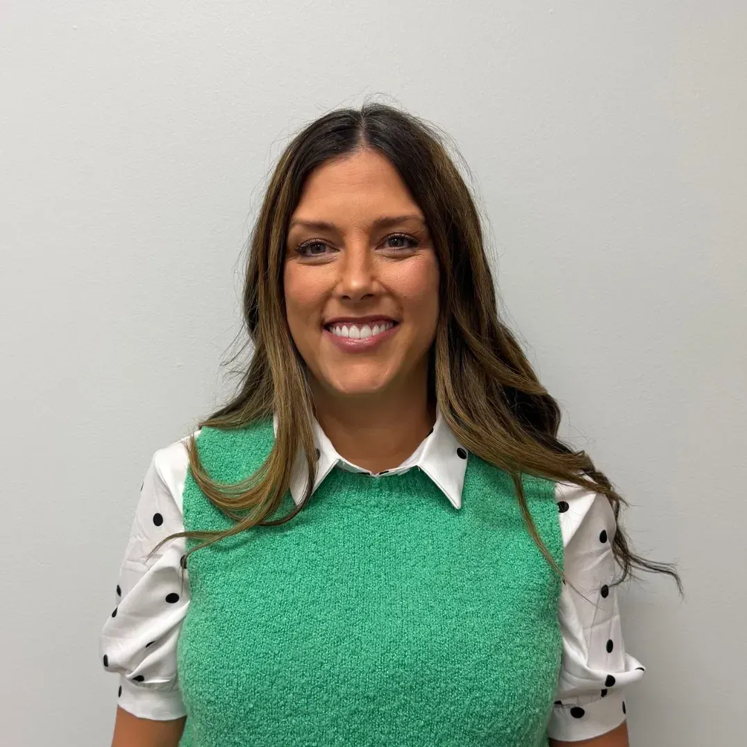 Woman with brown hair smiles, wearing a green vest over a white polka dot blouse, against a plain white wall.