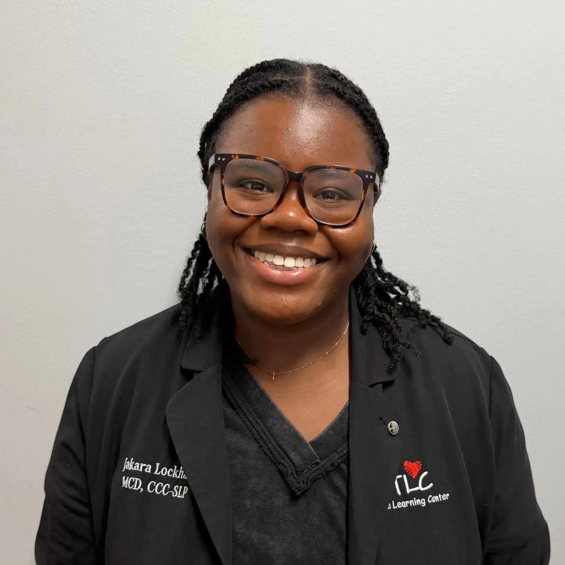 Woman wearing glasses, black jacket, and smiling, next to a logo, against a white wall.