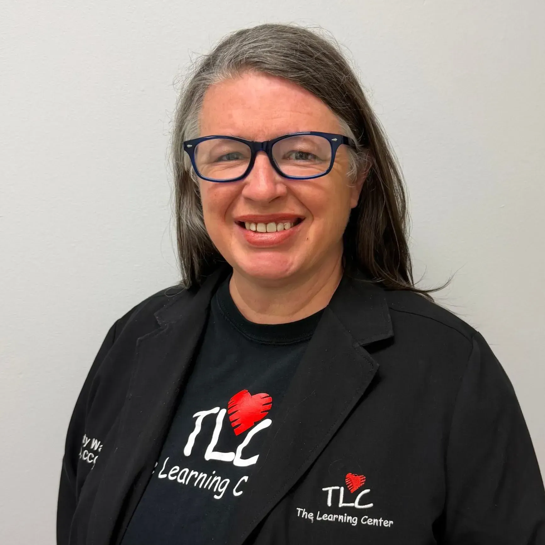 Woman wearing glasses and black jacket, smiling. T-shirt reads 