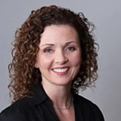 Woman with curly brown hair smiling, wearing a black shirt against a gray background.