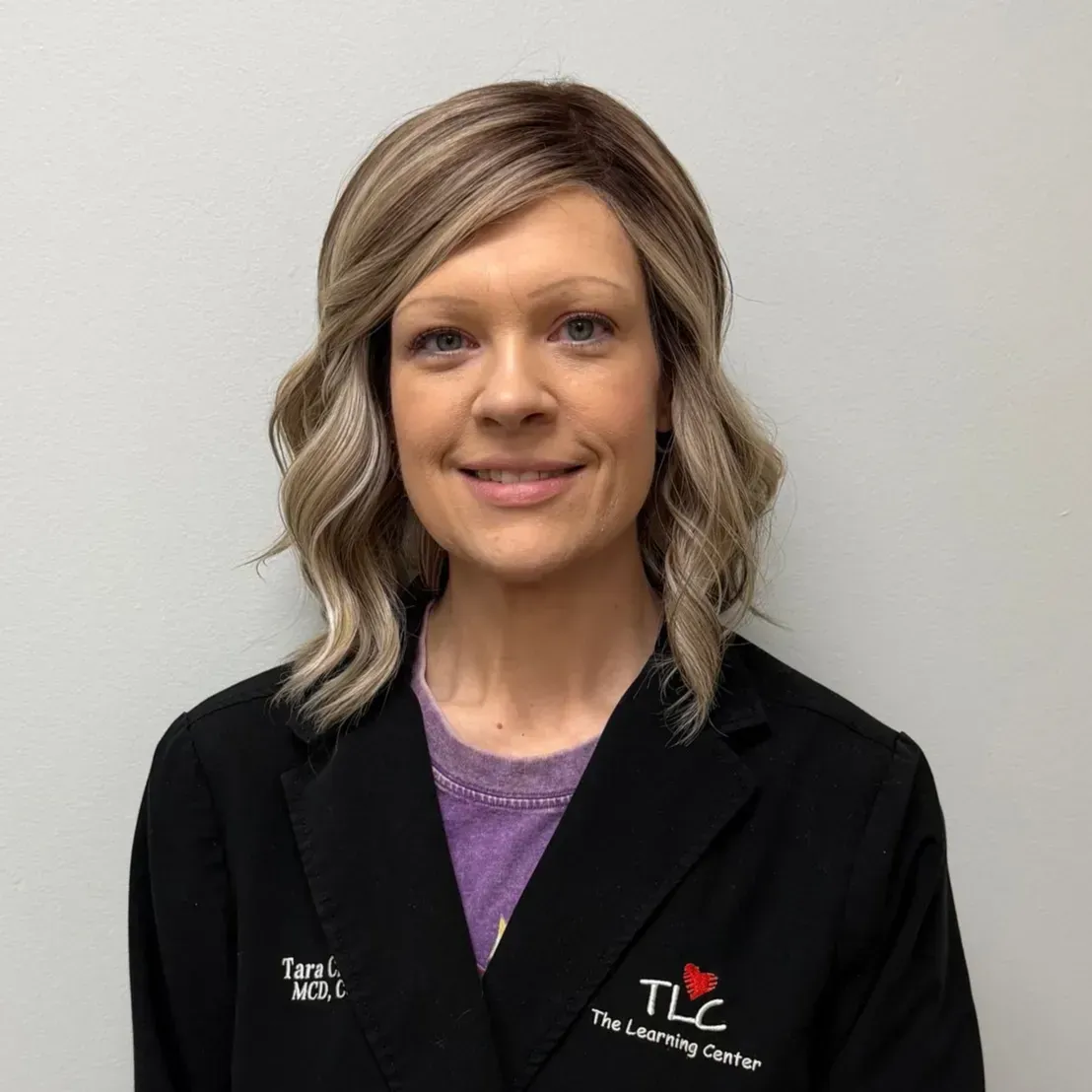 Woman in black jacket smiles, wearing light brown hair and a purple shirt. TLC logo on jacket.
