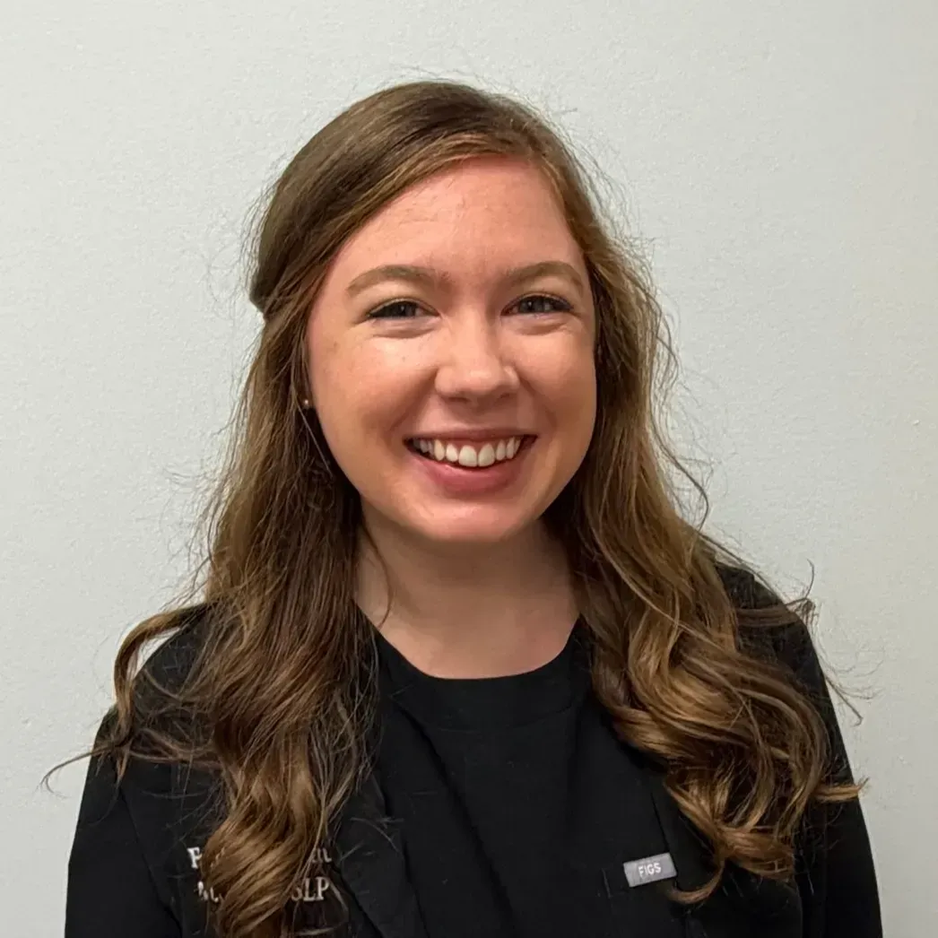 Woman with long, wavy brown hair, smiling, wearing a black shirt in front of a white wall.