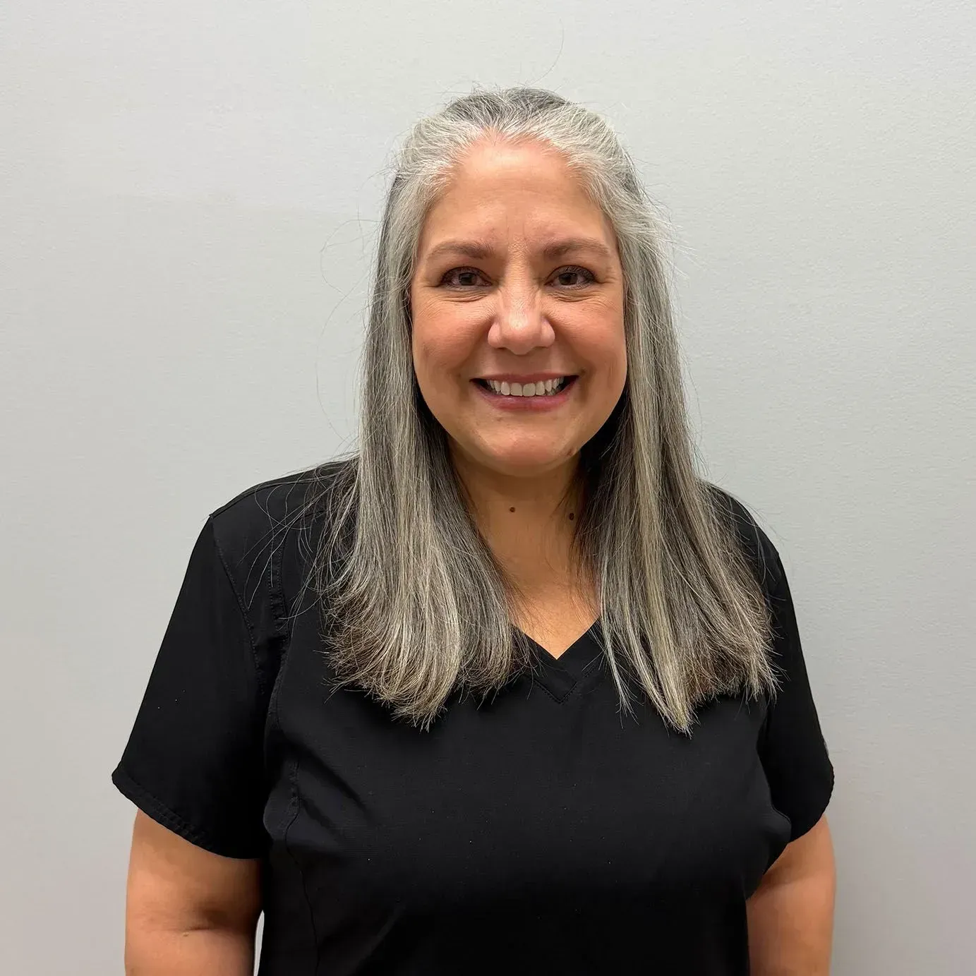 Woman with gray and dark hair wearing a black top smiles in front of a gray wall.