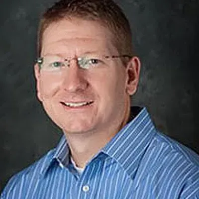 Man with glasses smiles, wearing blue striped shirt, looking at camera.