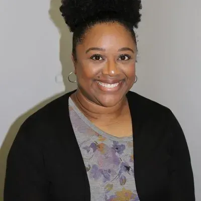 Woman with dark hair in bun smiles, wearing gray floral top and black cardigan.