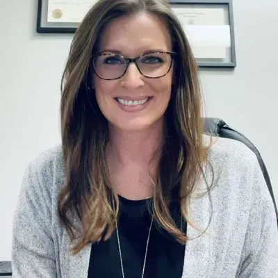 Woman with glasses smiles; wearing black shirt, gray sweater in an office.