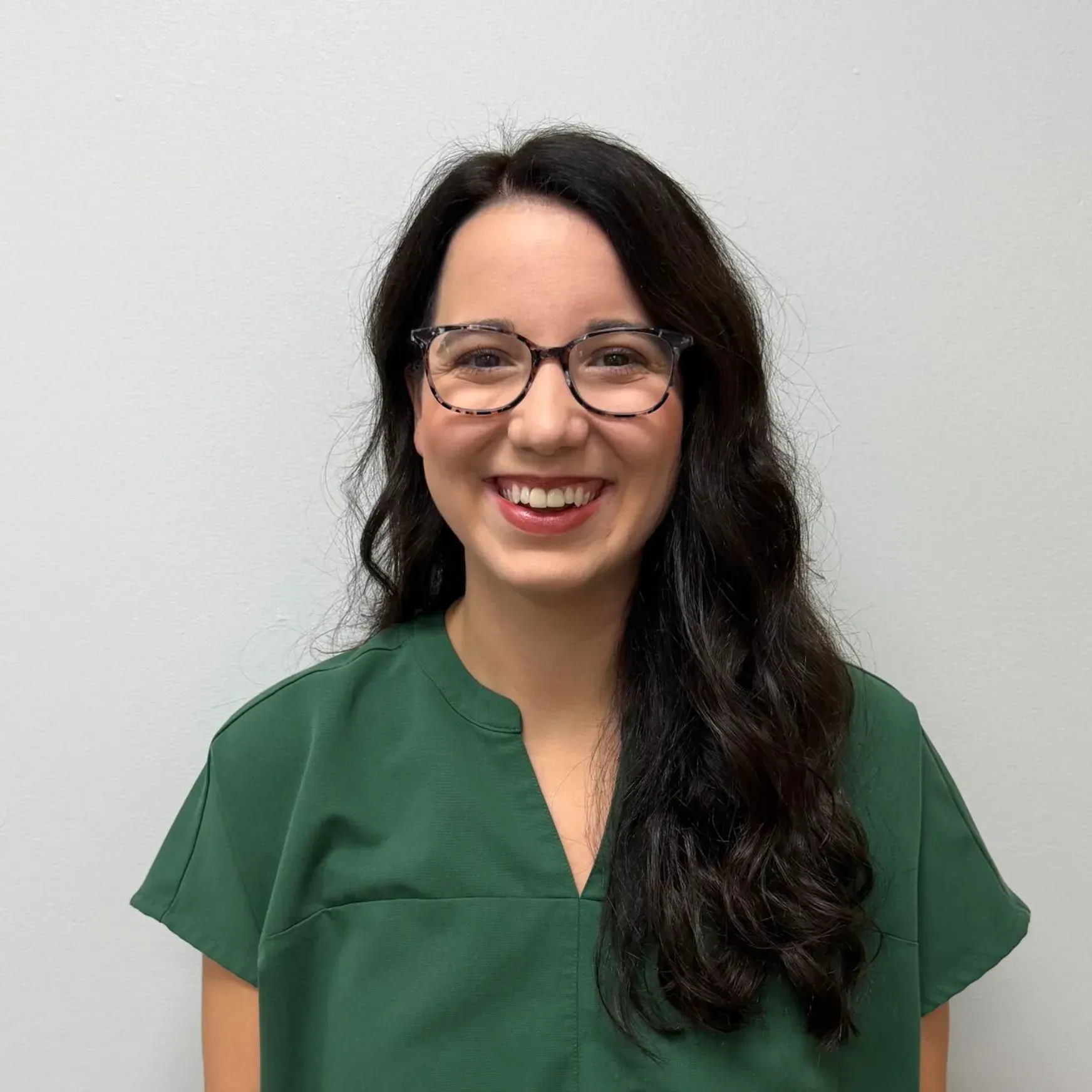 Woman with dark hair and glasses smiles, wearing a green top, against a white wall.