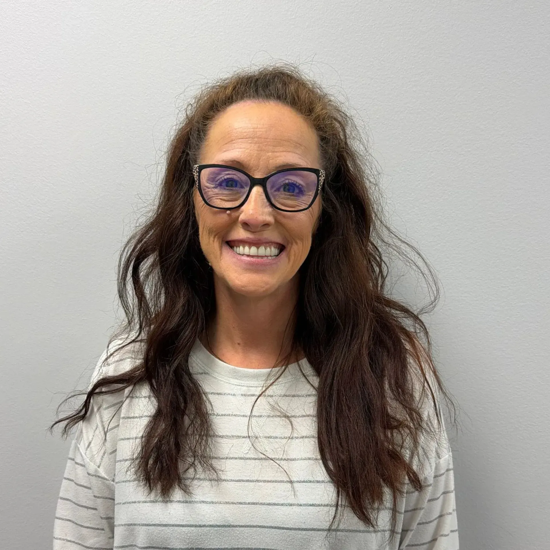 Woman with glasses smiles, wearing striped shirt, against a white wall.