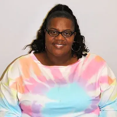 Woman wearing glasses and a tie-dye shirt smiles. Her hair is pulled back in a high ponytail.