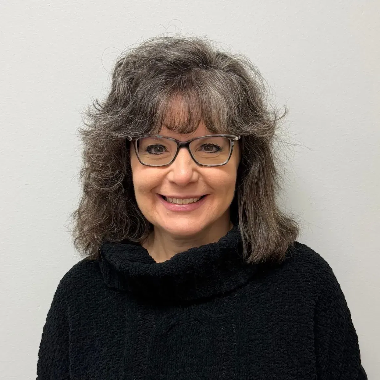Woman wearing glasses and black sweater smiles against a white background.