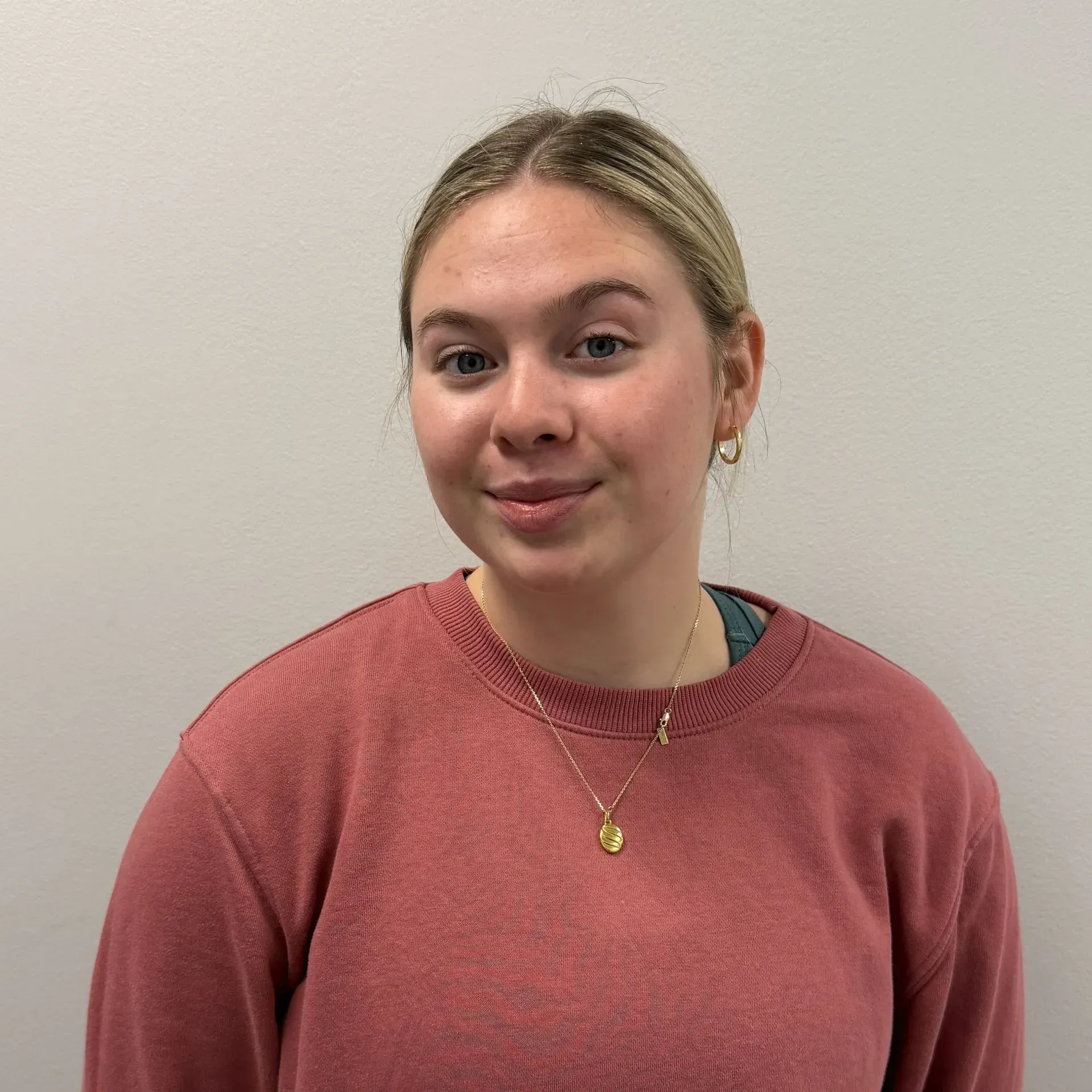 Woman with blonde hair wearing a pink sweater, gold necklace, and earrings smiling at the camera against a white wall.