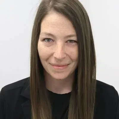 Woman with long brown hair, wearing a black blazer, smiles at the camera. Neutral background.