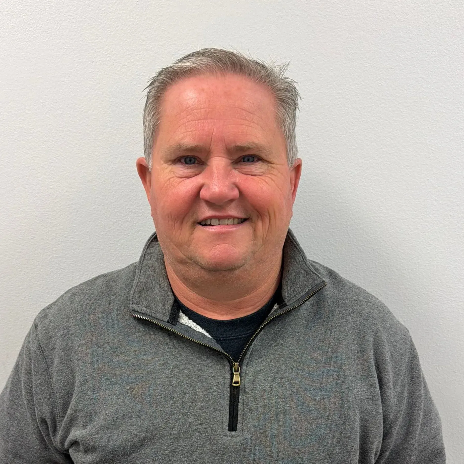 Man in gray zip-up sweater smiles in front of a white wall.