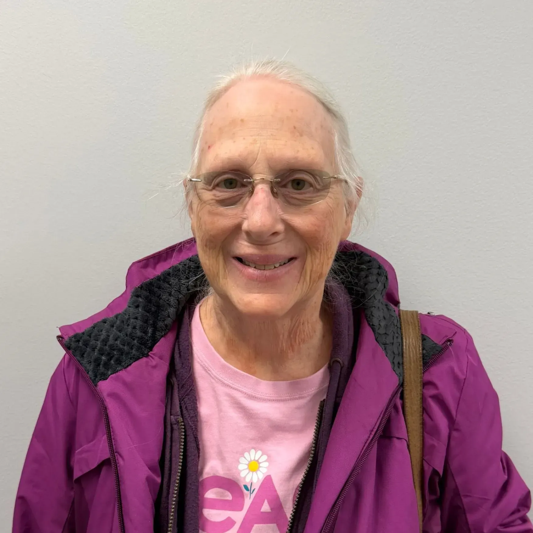 Woman with glasses and pink shirt, wearing a purple jacket, smiling. Against a white wall.