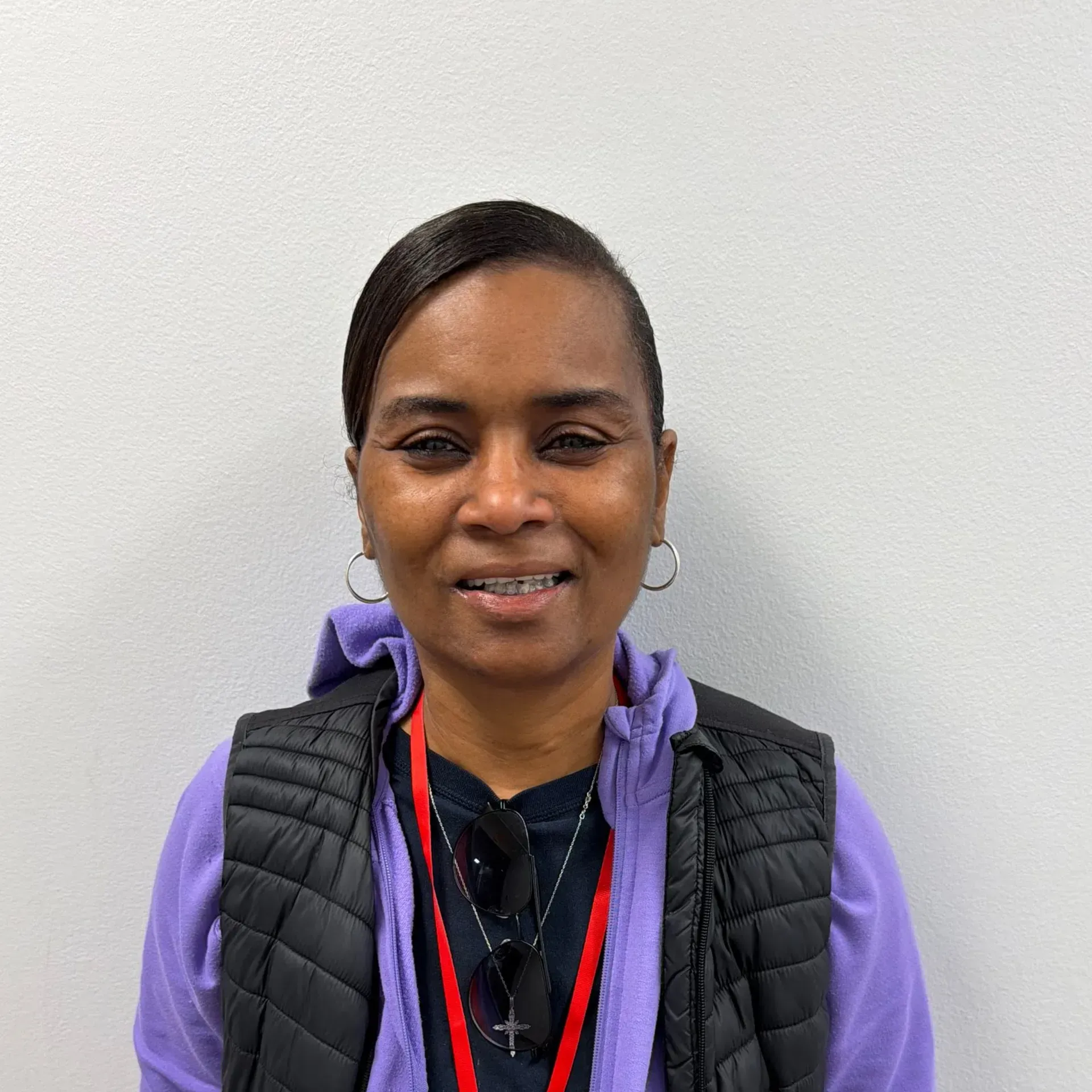 Woman wearing a black vest and purple hoodie smiles, standing in front of a white wall.