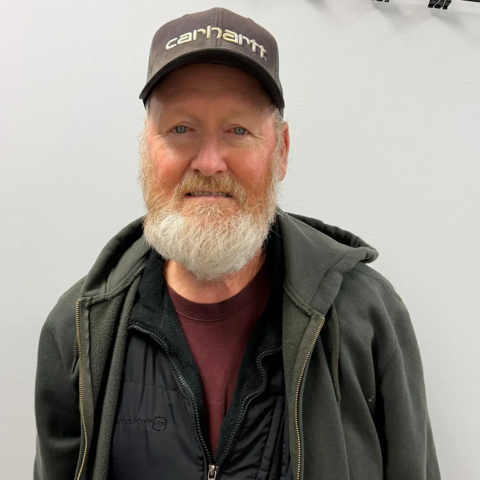 Man with white beard, wearing a Carhartt hat and hooded jacket, smiling.