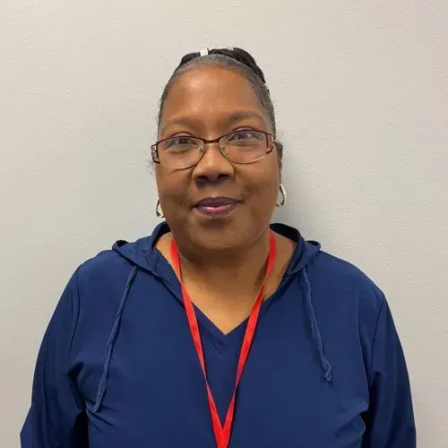 Woman wearing glasses and a blue top with a red lanyard, standing in front of a white wall.