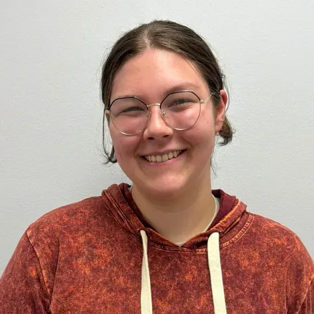 Woman with glasses smiles, wearing an orange hoodie, against a plain background.