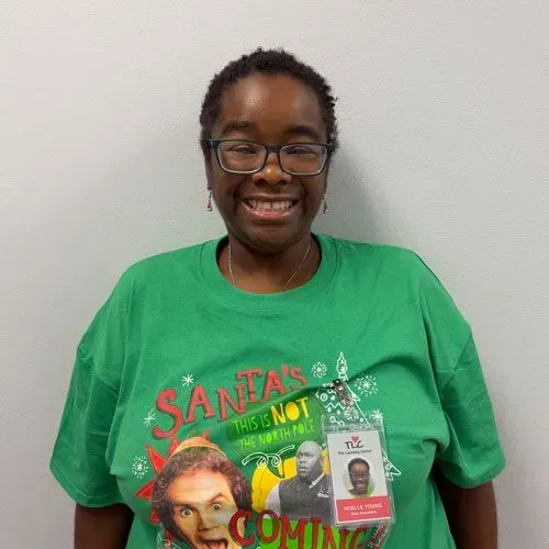Woman wearing glasses and a green Christmas shirt smiles, with an ID badge clipped to her shirt.