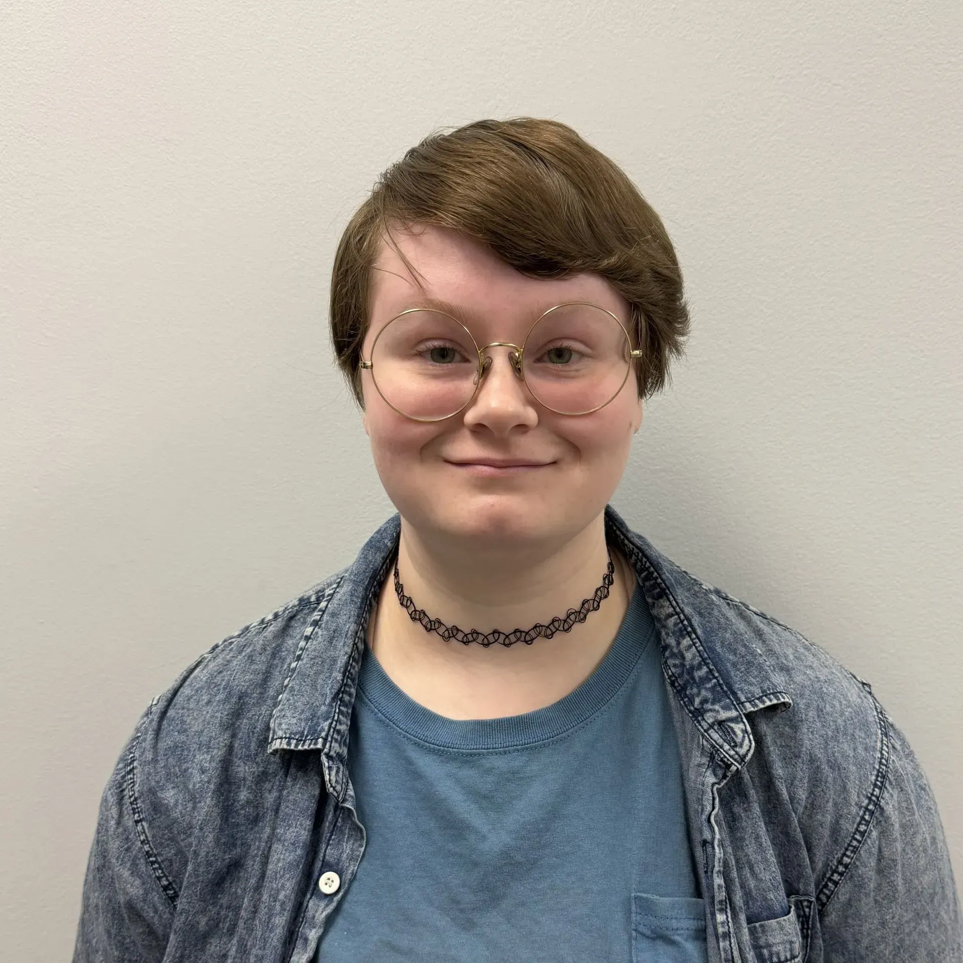 Person with glasses and short brown hair, wearing a denim jacket and a choker, smiling.