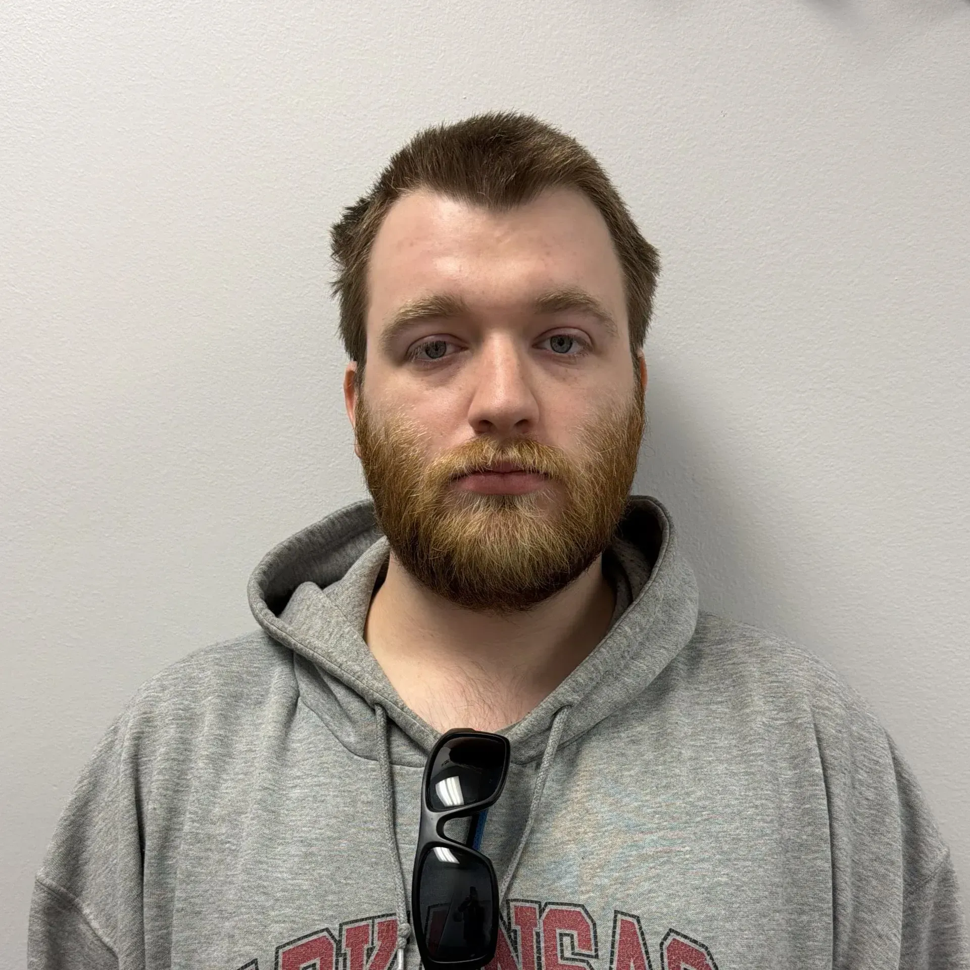 Man with a beard, wearing a gray hoodie and sunglasses, stands in front of a white wall.