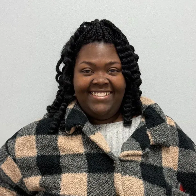 Woman with braided hair, smiling, wearing a black and beige plaid coat, in front of a white wall.