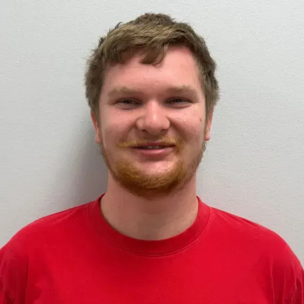 Man with reddish-brown hair and beard smiles, wearing a red t-shirt in front of a white wall.