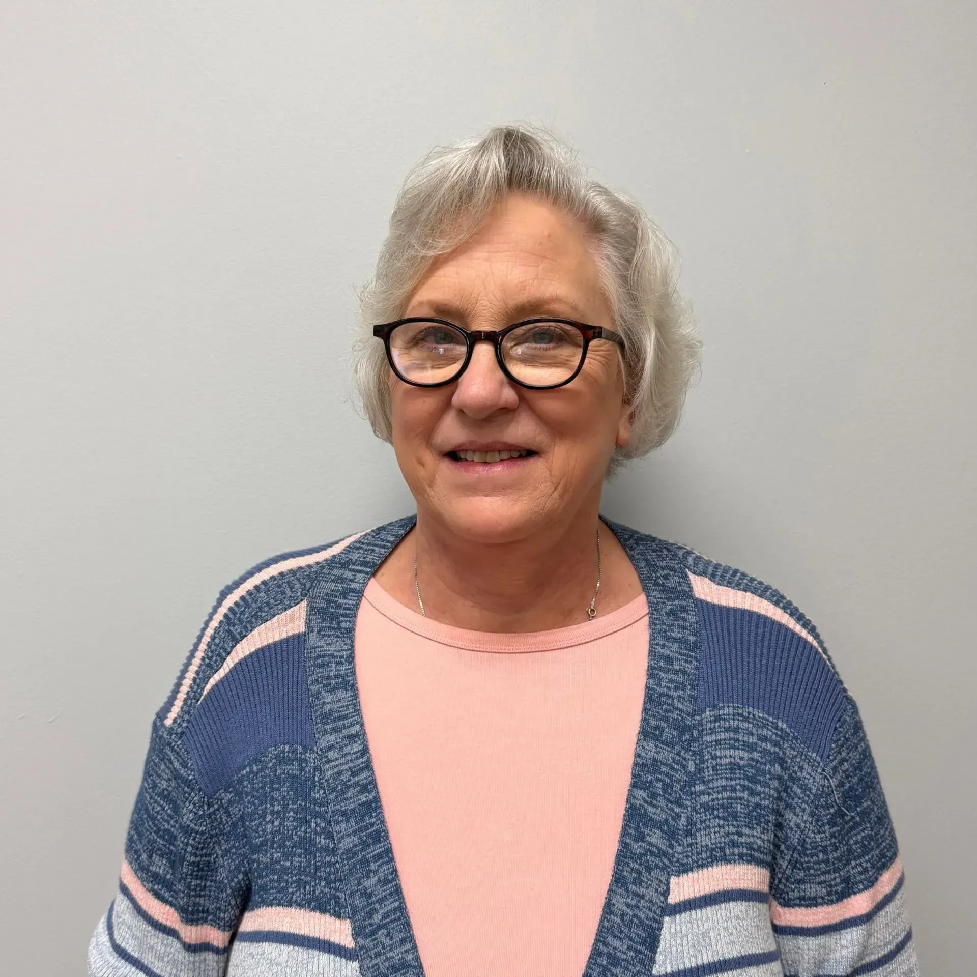 Woman wearing glasses, a pink shirt, and a blue sweater smiles in front of a gray wall.