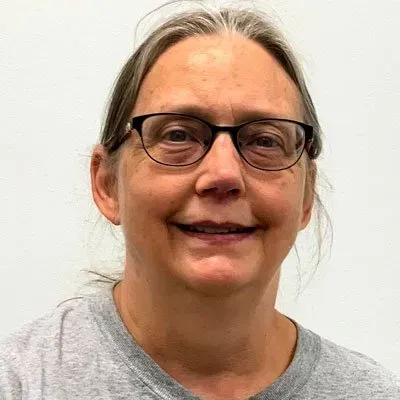 Woman with glasses smiling, wearing a gray shirt, in front of a white wall.