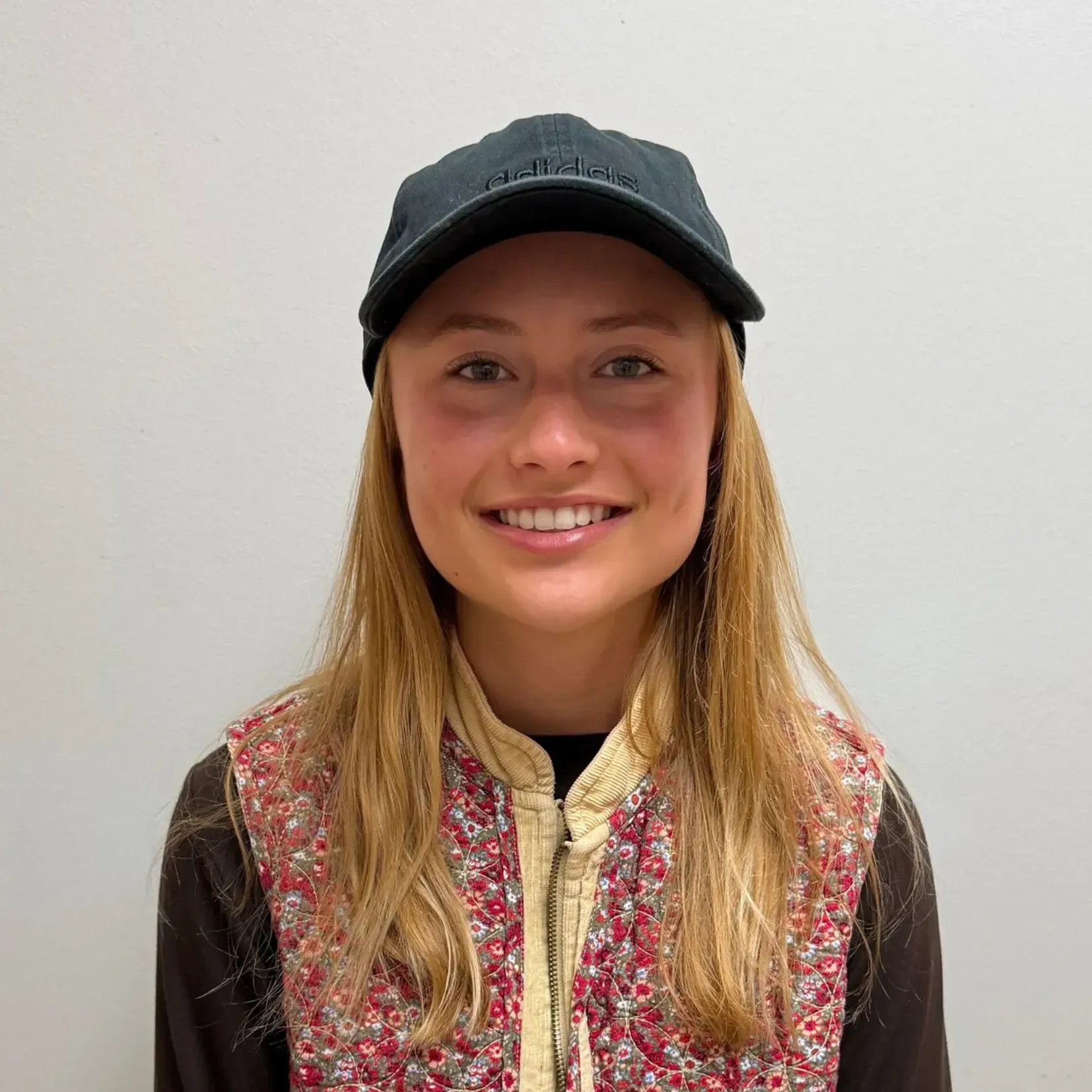 Woman with blonde hair wearing a black cap and floral vest smiles at the camera.