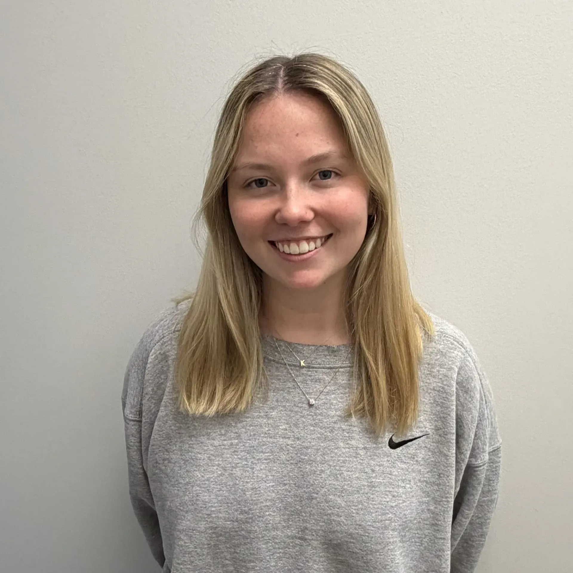 Blonde person smiling, wearing a gray sweatshirt, standing in front of a light wall.