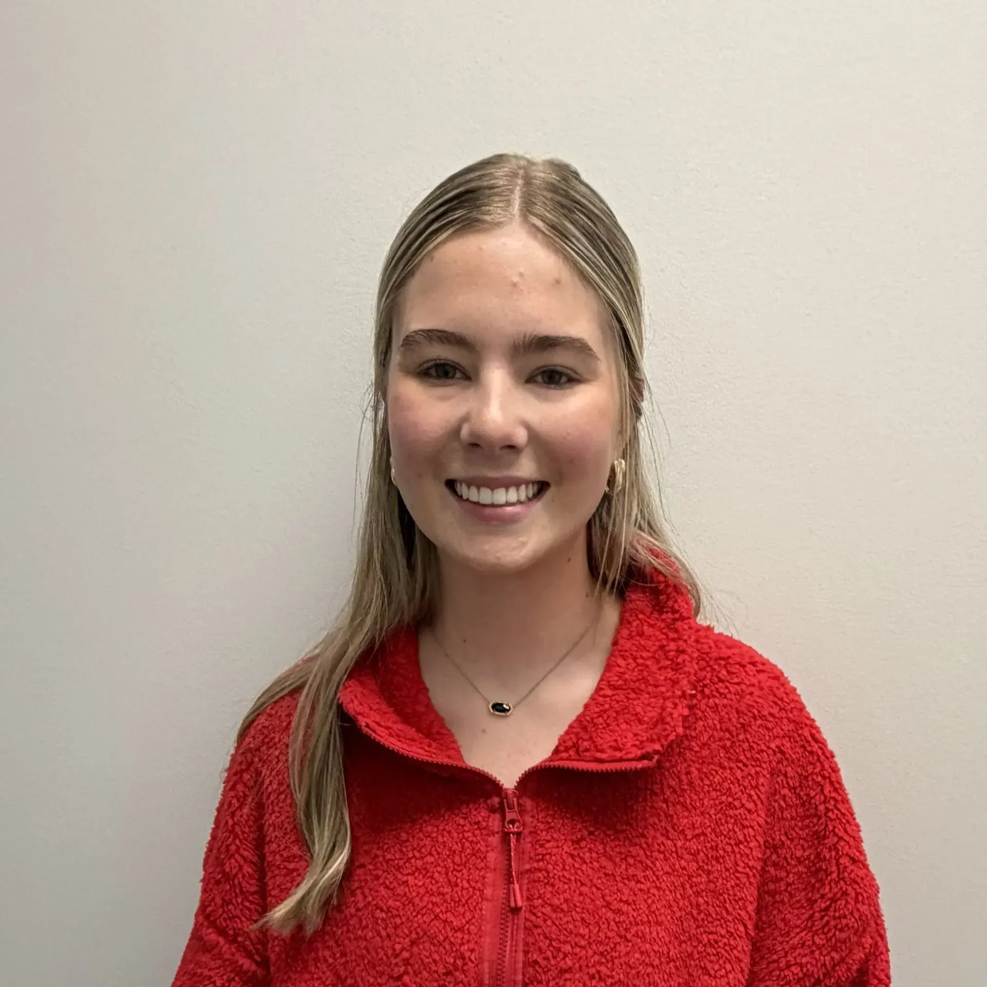 Woman with blonde hair wearing a red zip-up fleece, smiling against a white wall.