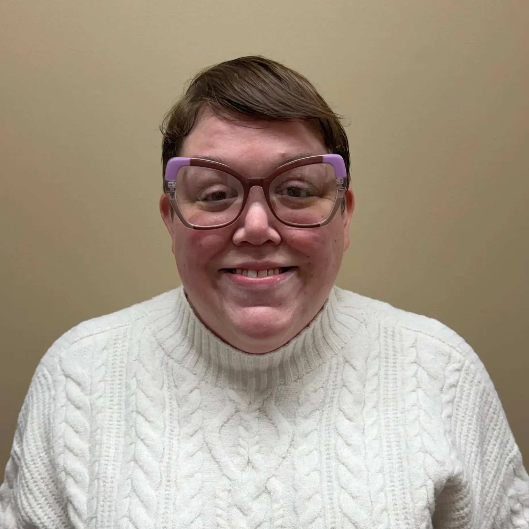 Woman with glasses smiles wearing a white sweater against a beige wall.