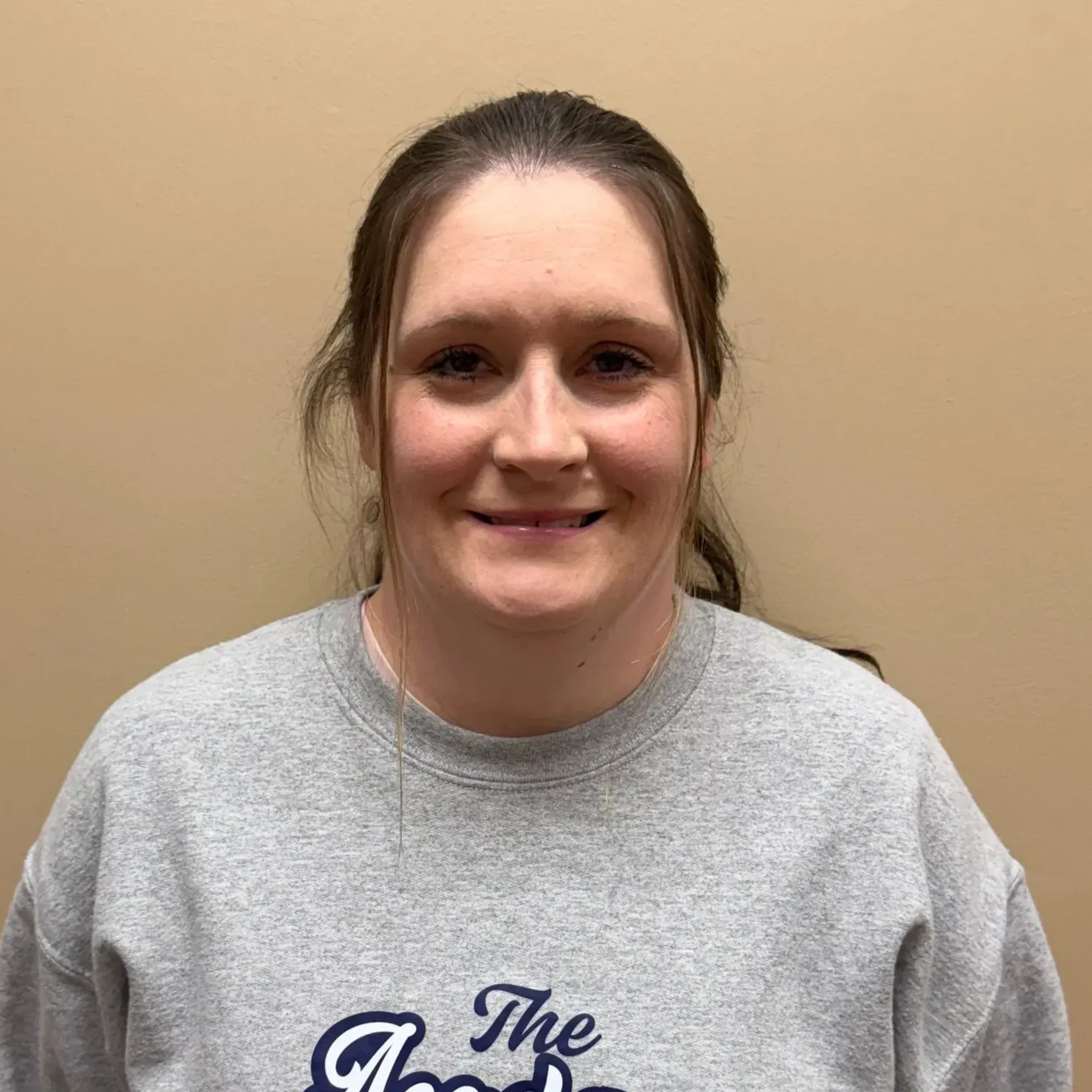 Woman in a gray sweatshirt smiles, looking at the camera against a beige wall.