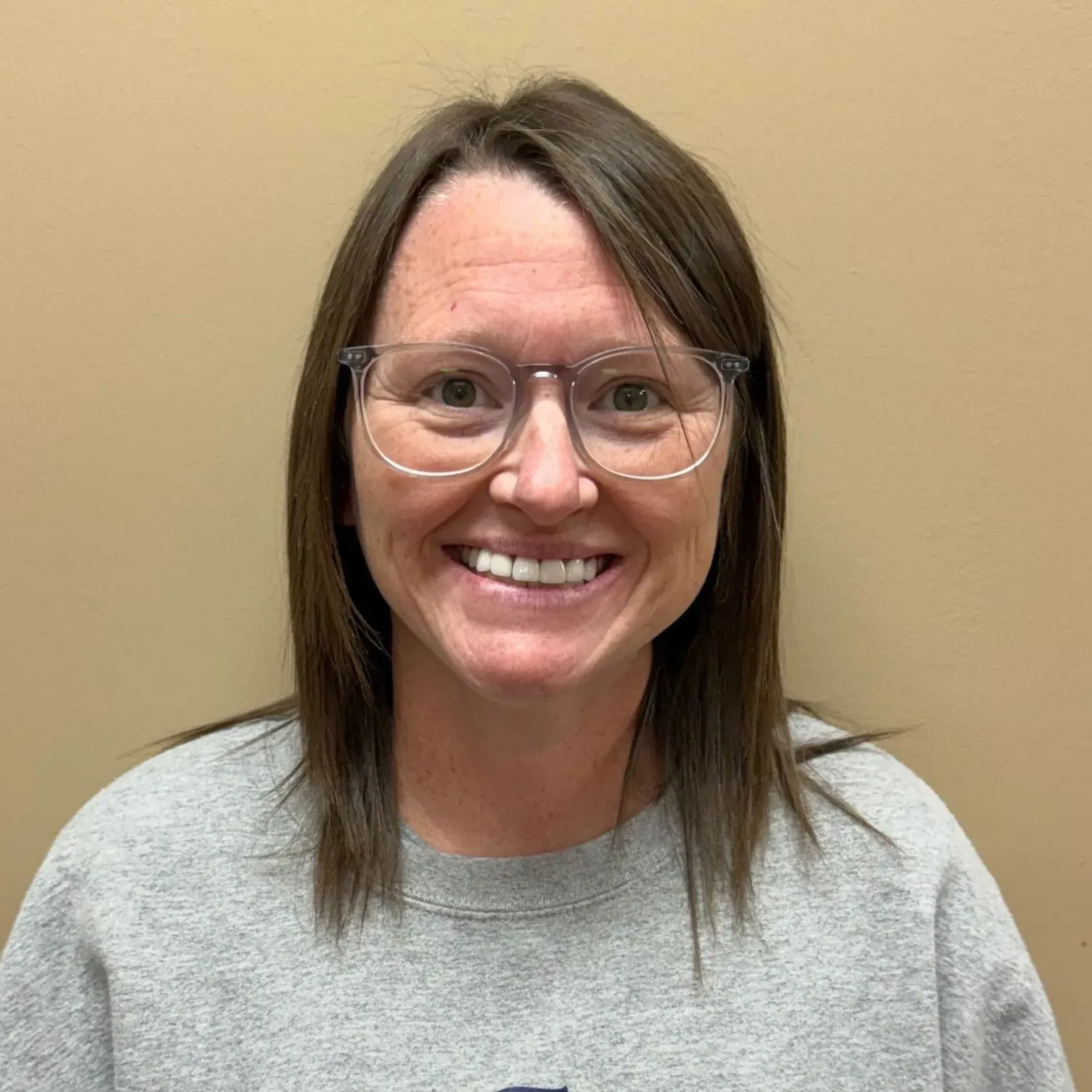 Woman with glasses smiles at the camera, wearing a gray sweatshirt, in front of a tan wall.