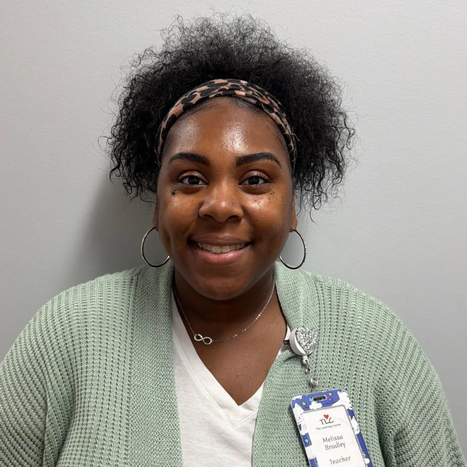 Woman with dark curly hair wearing a leopard print headband, hoop earrings, and a light green cardigan smiles at the camera.