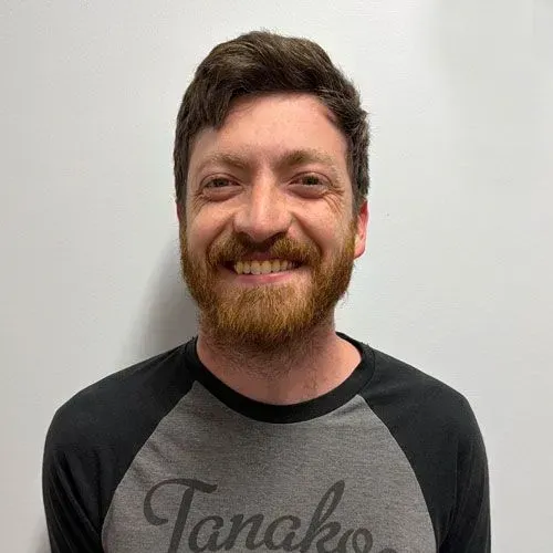 Man with brown hair and beard smiles, wearing a gray and black raglan shirt in front of a white wall.