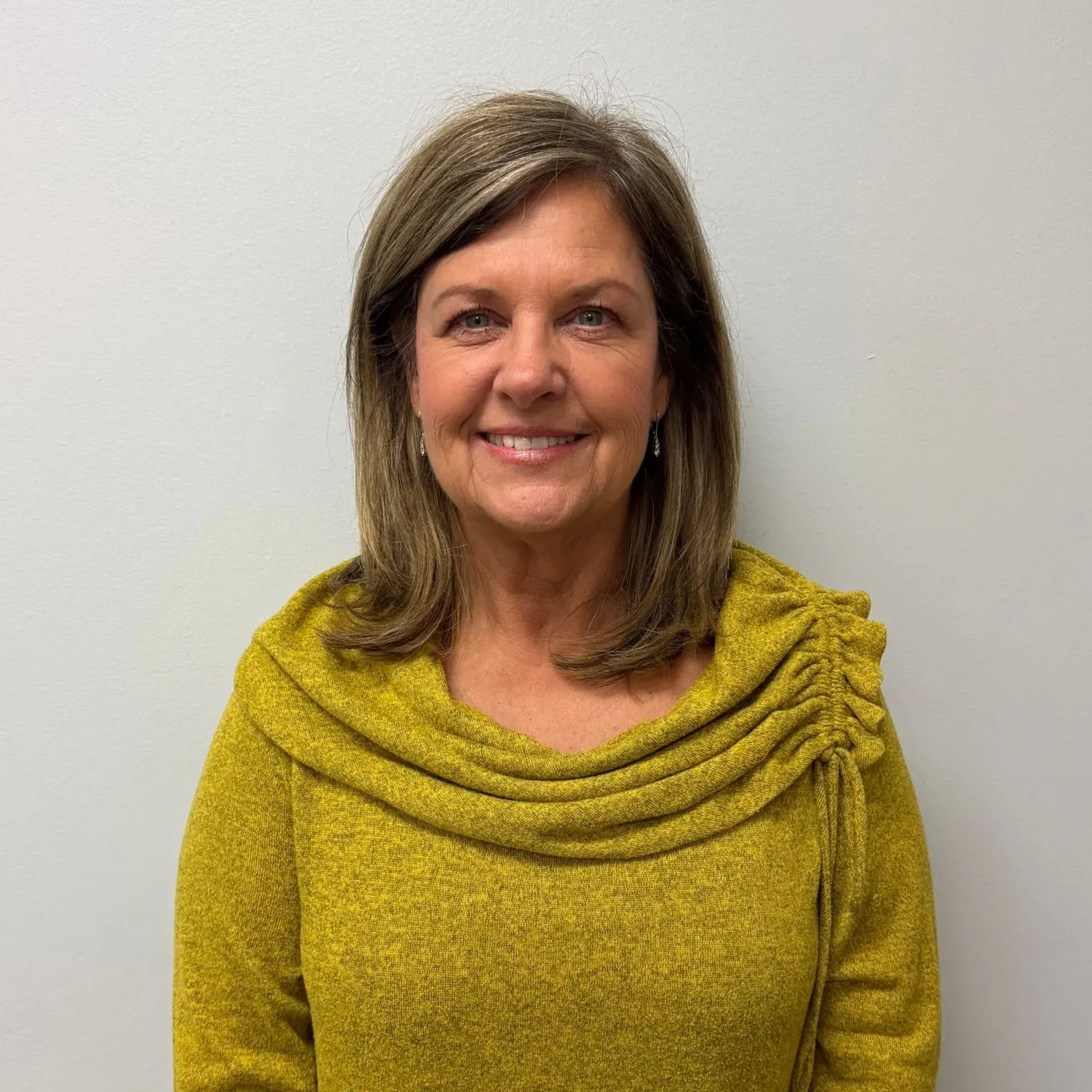 Woman with shoulder-length brown hair, wearing a mustard-yellow sweater, smiling, against a plain white wall.