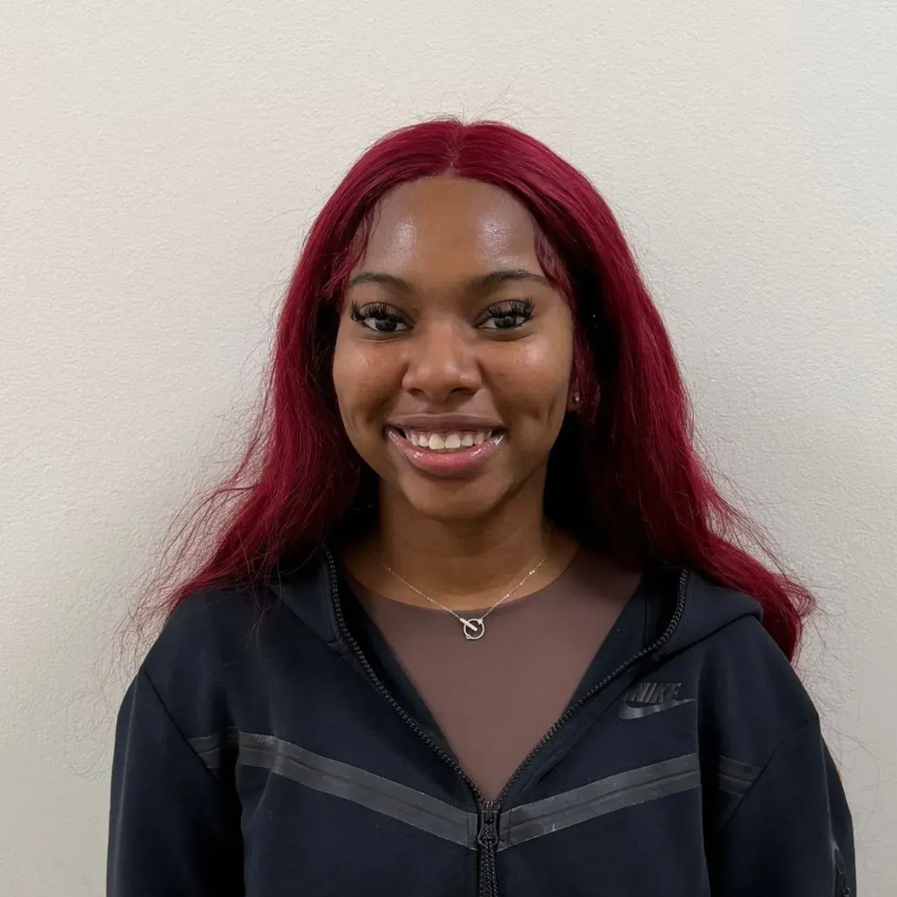 Person with burgundy hair smiling, wearing a black zip-up jacket over a brown top. Light background.