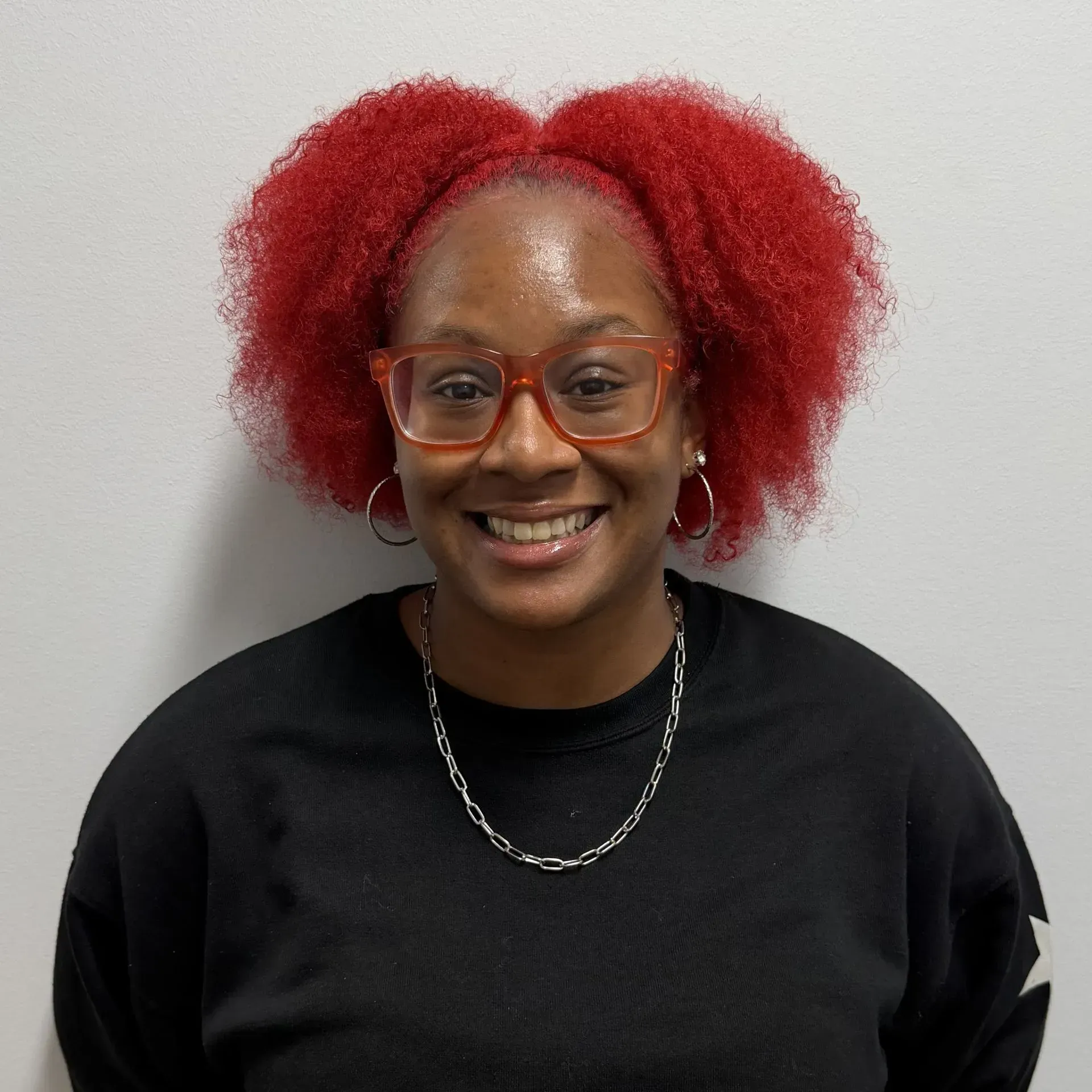 Woman with red hair and glasses smiles, wearing a black sweatshirt and silver necklace.