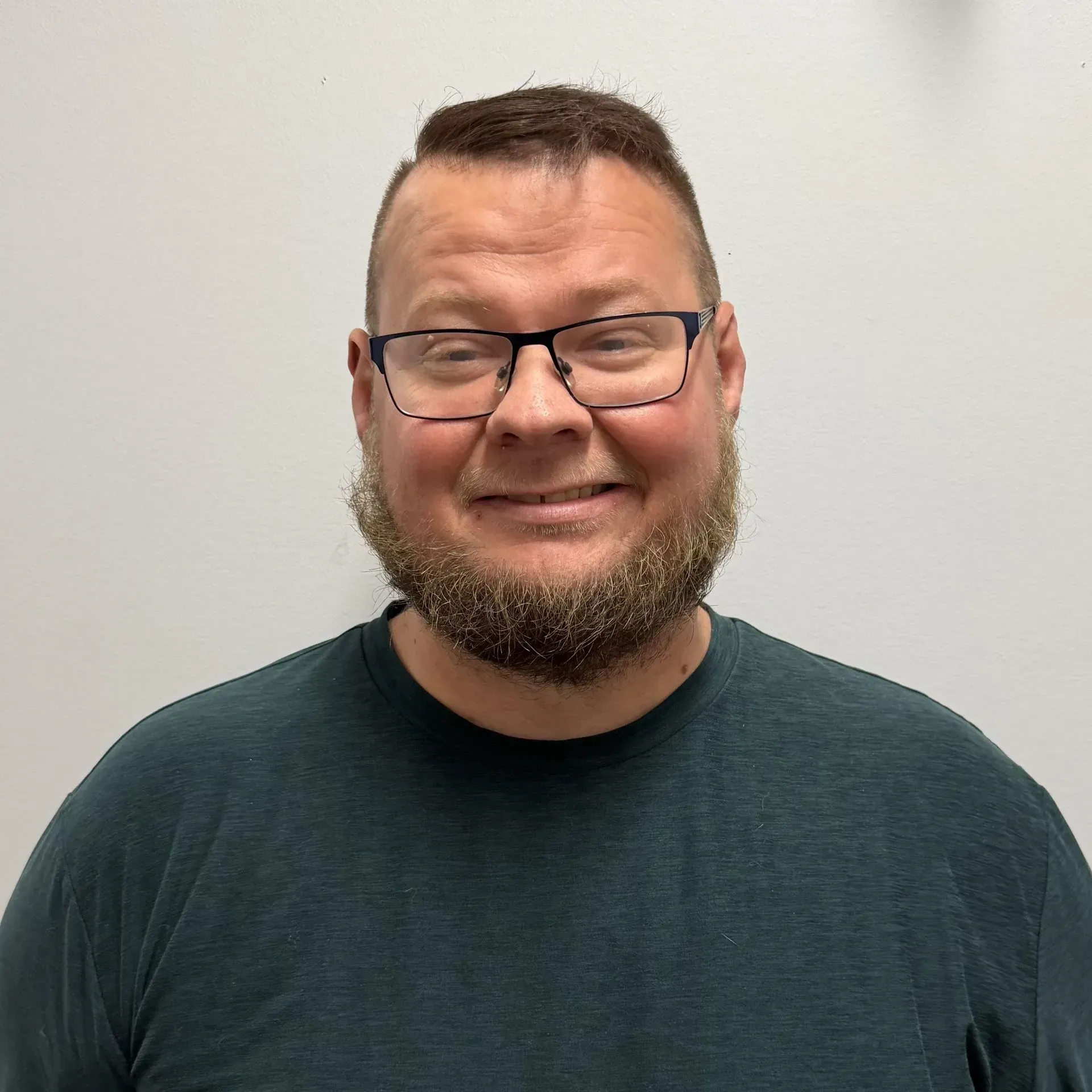 Man with a beard and glasses smiling, wearing a dark green shirt, in front of a white wall.