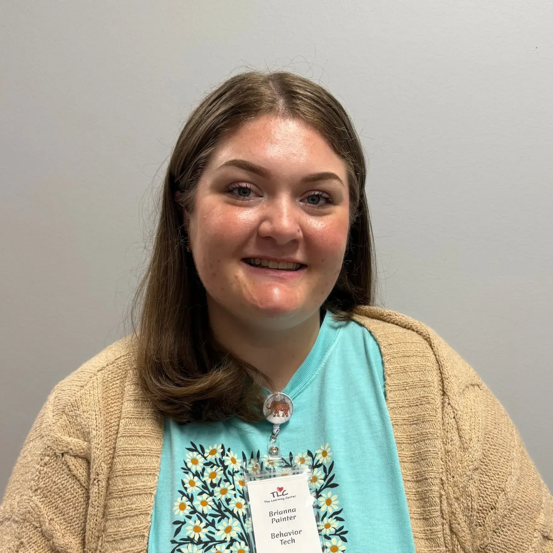 Woman in a blue shirt and tan sweater smiles at the camera, wearing an ID badge.