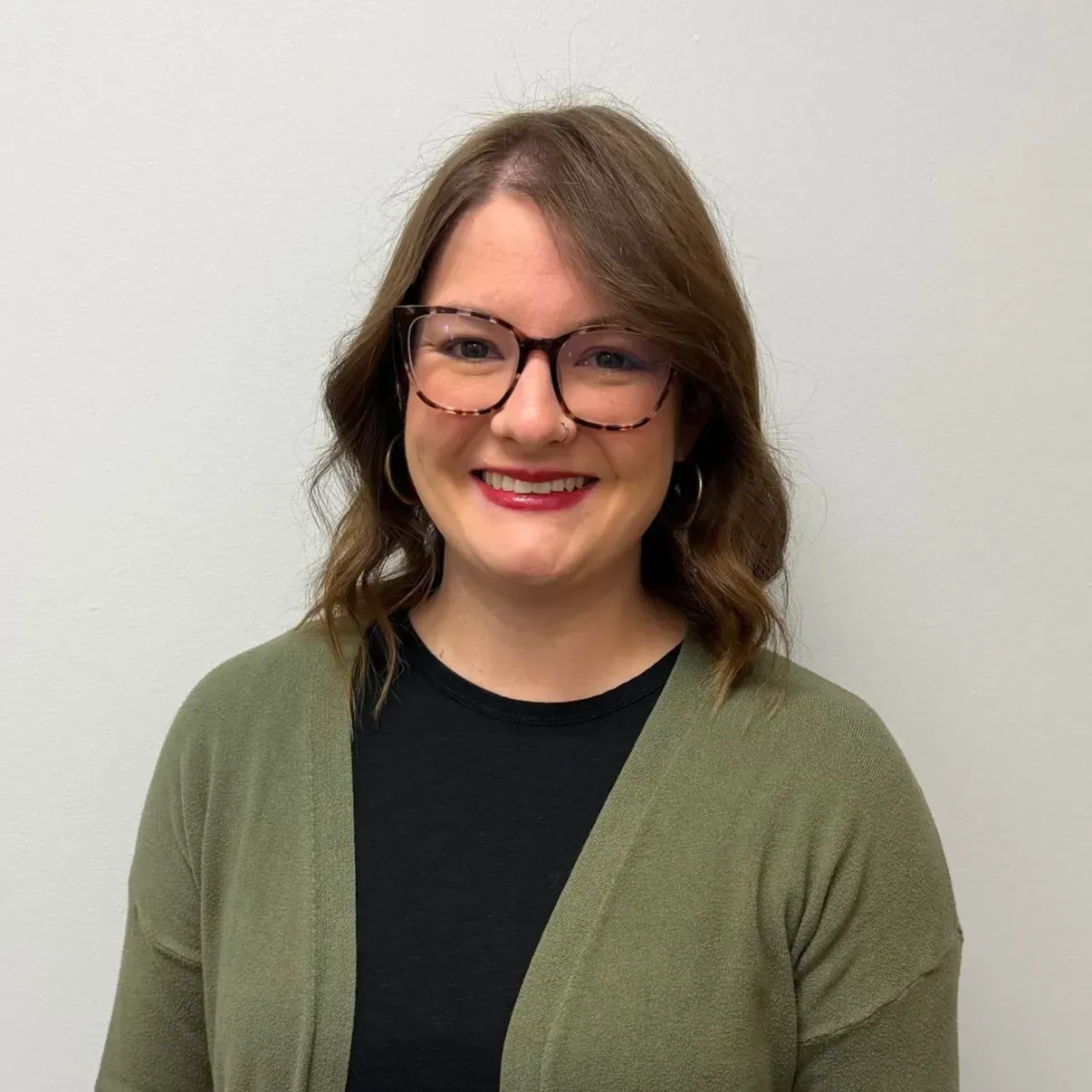Woman with glasses smiles wearing a green cardigan and black shirt in front of a white wall.