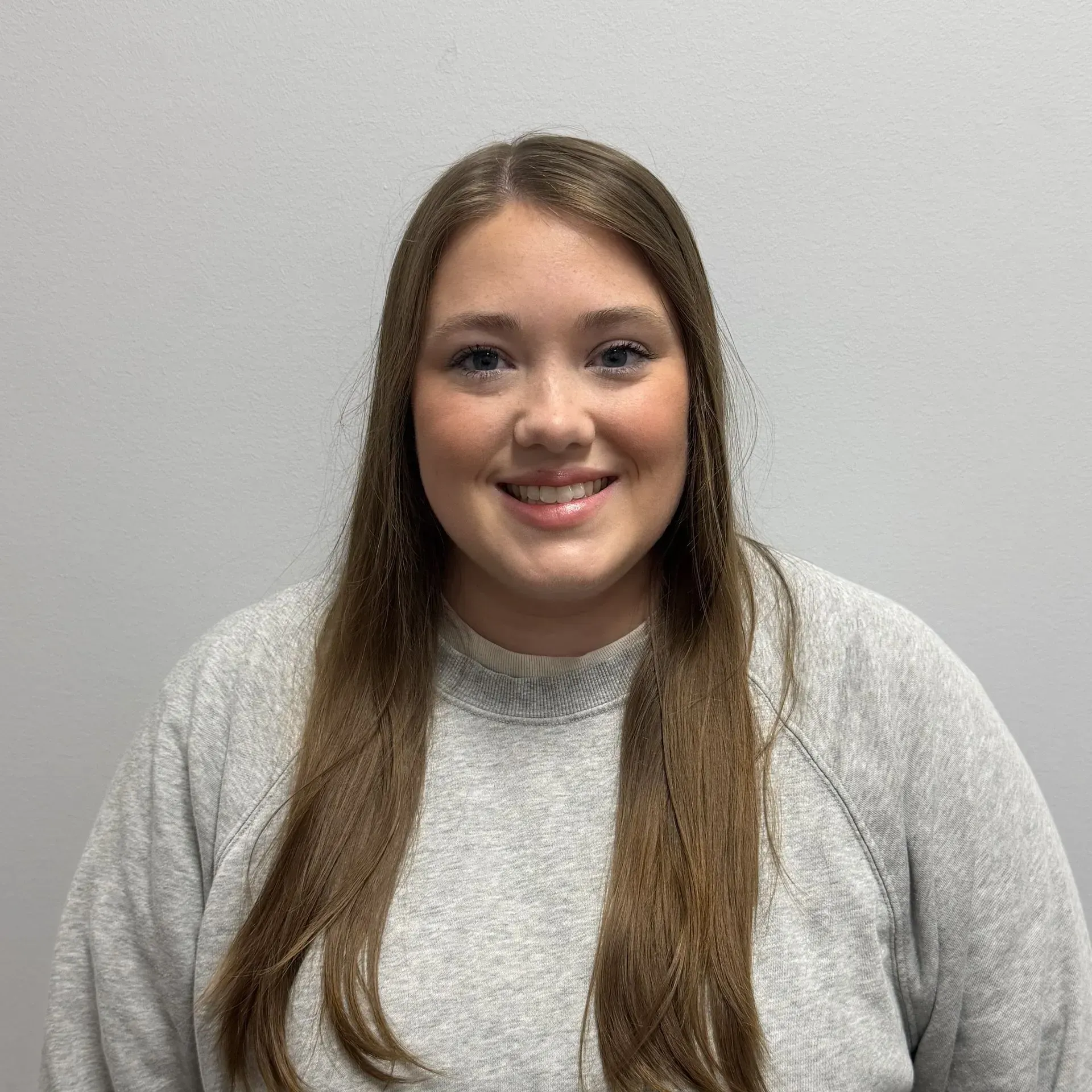 Woman with long brown hair smiles, wearing a gray sweater, standing in front of a gray wall.