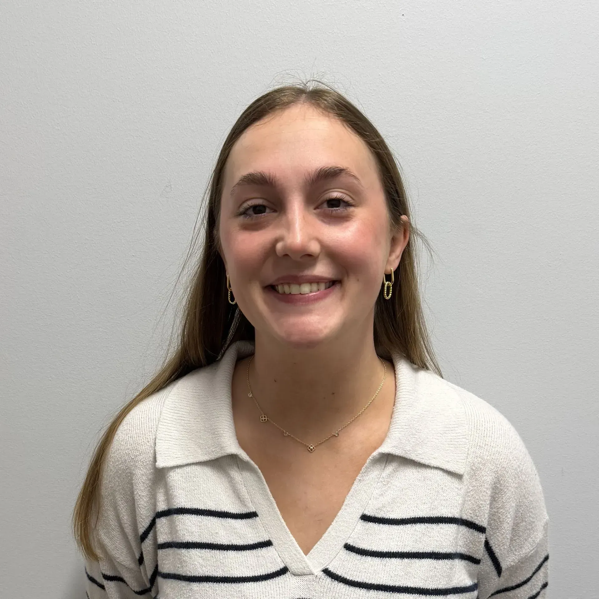 Woman with long brown hair smiling, wearing a cream sweater with black stripes, and gold jewelry.
