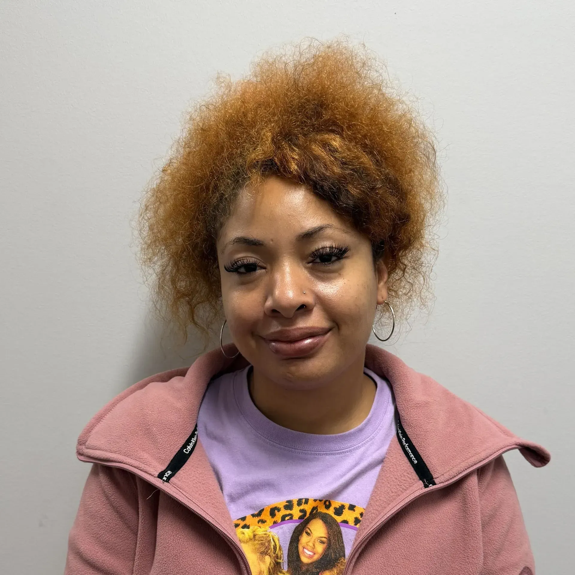 Woman with curly orange hair smiles, wearing pink jacket and purple t-shirt.