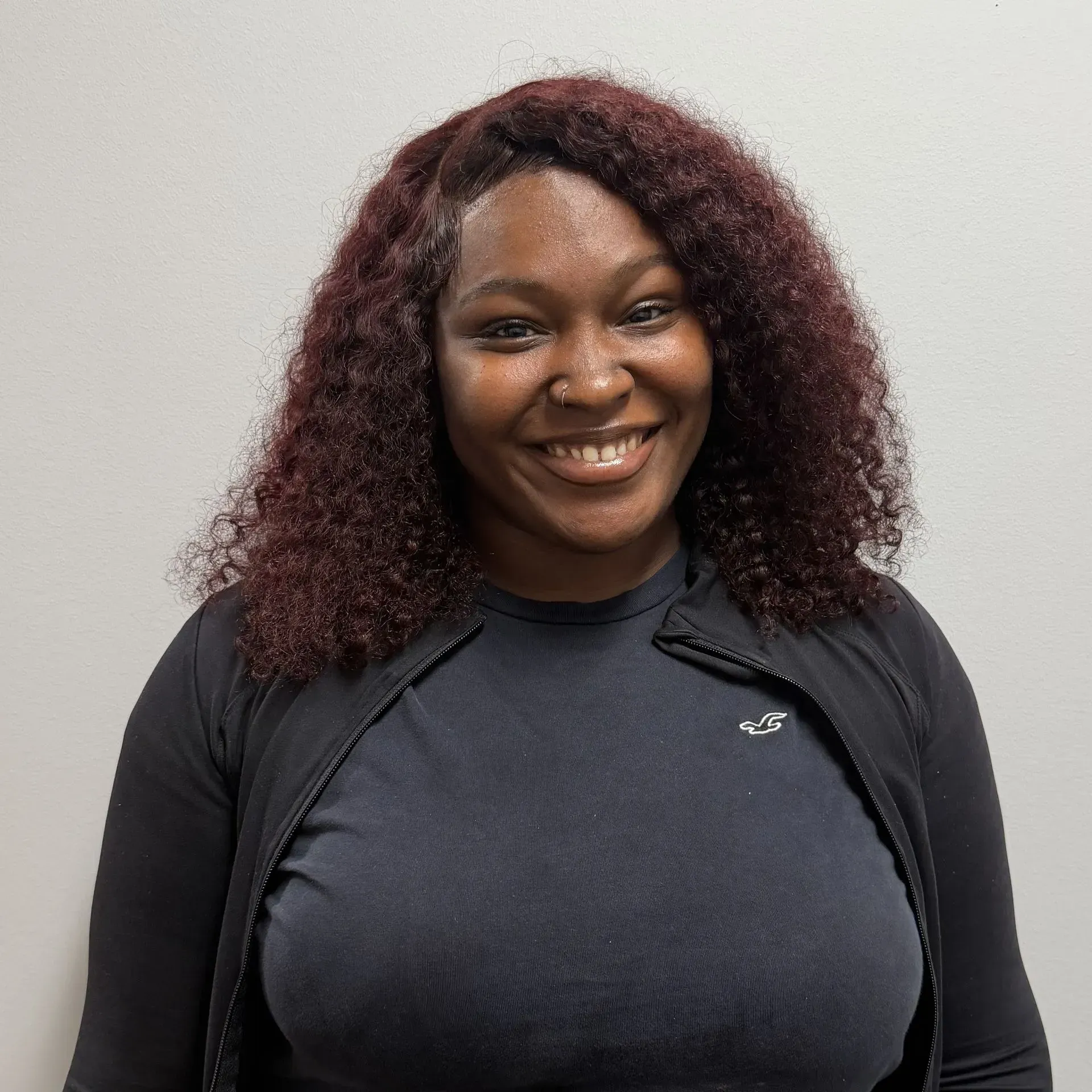 Woman with curly maroon hair smiles, wearing a black shirt and jacket, against a white wall.