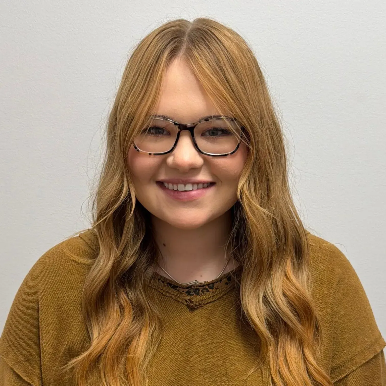 Woman with long, wavy blonde hair, glasses, and a brown sweater smiles at the camera against a plain backdrop.