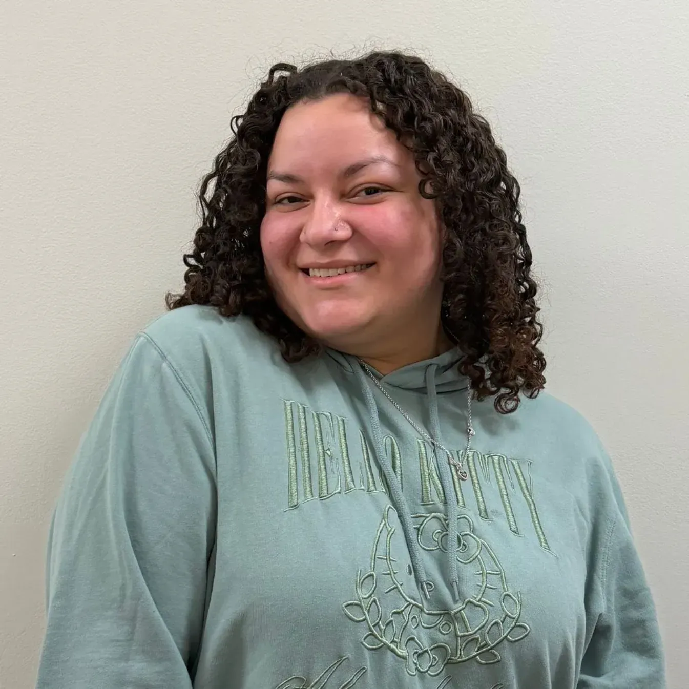 Woman with curly hair smiles, wearing a light blue sweatshirt with embroidered design, against a plain wall.